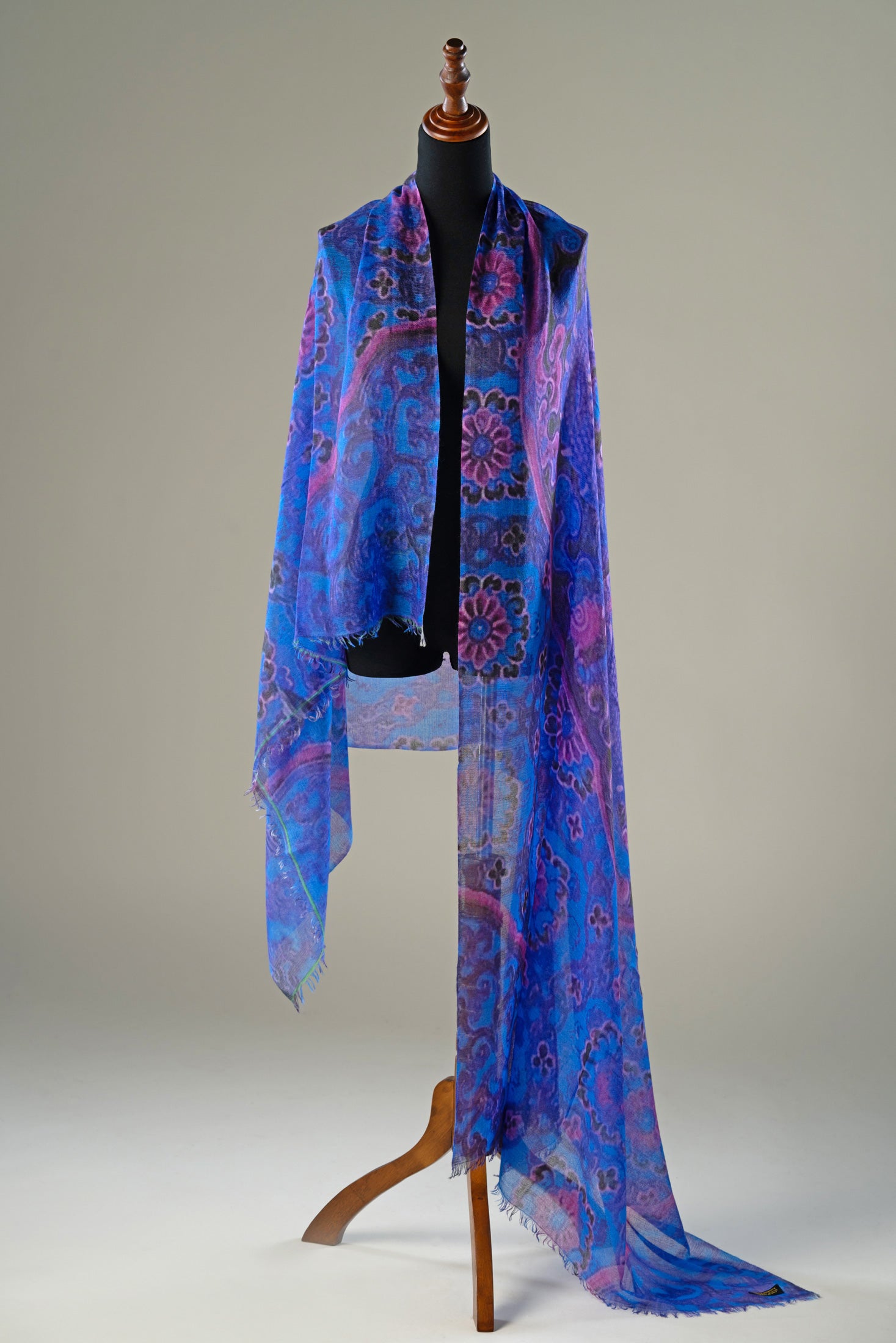 A lightweight blue scarf with a purple and pink paisley pattern draped over a black dress form mannequin on a wooden stand, set against a neutral gray studio background.