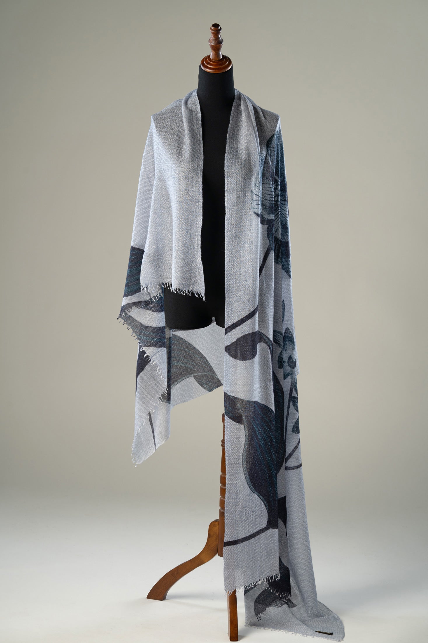 Luxury handwoven grey cashmere shawl featuring abstract monochrome botanical motifs and delicate fringed edges, elegantly displayed on a wooden stand.
