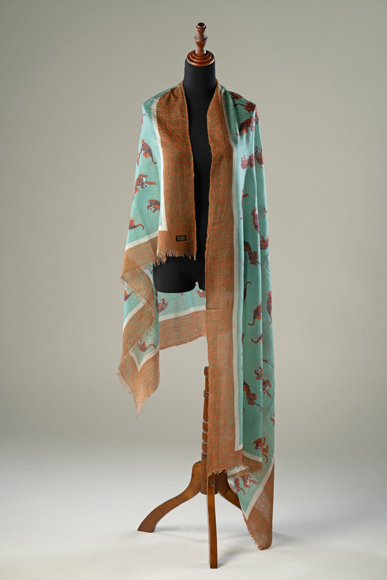 Luxury handwoven cashmere shawl in aqua green featuring artistic terracotta tiger motifs, intricate geometric borders, and delicate fringed edges elegantly displayed on a wooden stand.